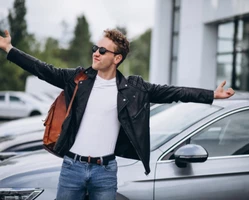 Парень в черной кожаной куртке с коричневым рюкзаком и очках стоит раскрыв руки на фоне серого автомобиля