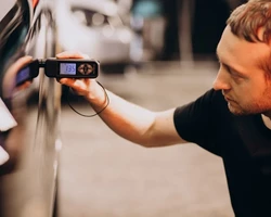 мужчина в черной футболке держит в руке толщиномер и проверяет толщину краски на автомобиле