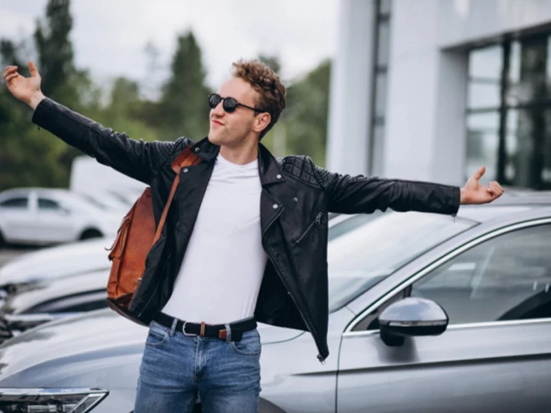 Парень в черной кожаной куртке с коричневым рюкзаком и очках стоит раскрыв руки на фоне серого автомобиля