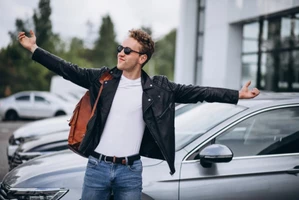 Парень в черной кожаной куртке с коричневым рюкзаком и очках стоит раскрыв руки на фоне серого автомобиля