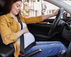 беременная женщина в белом свитере сидит за рулем автомобиля пристегнутая ремнем безопасности